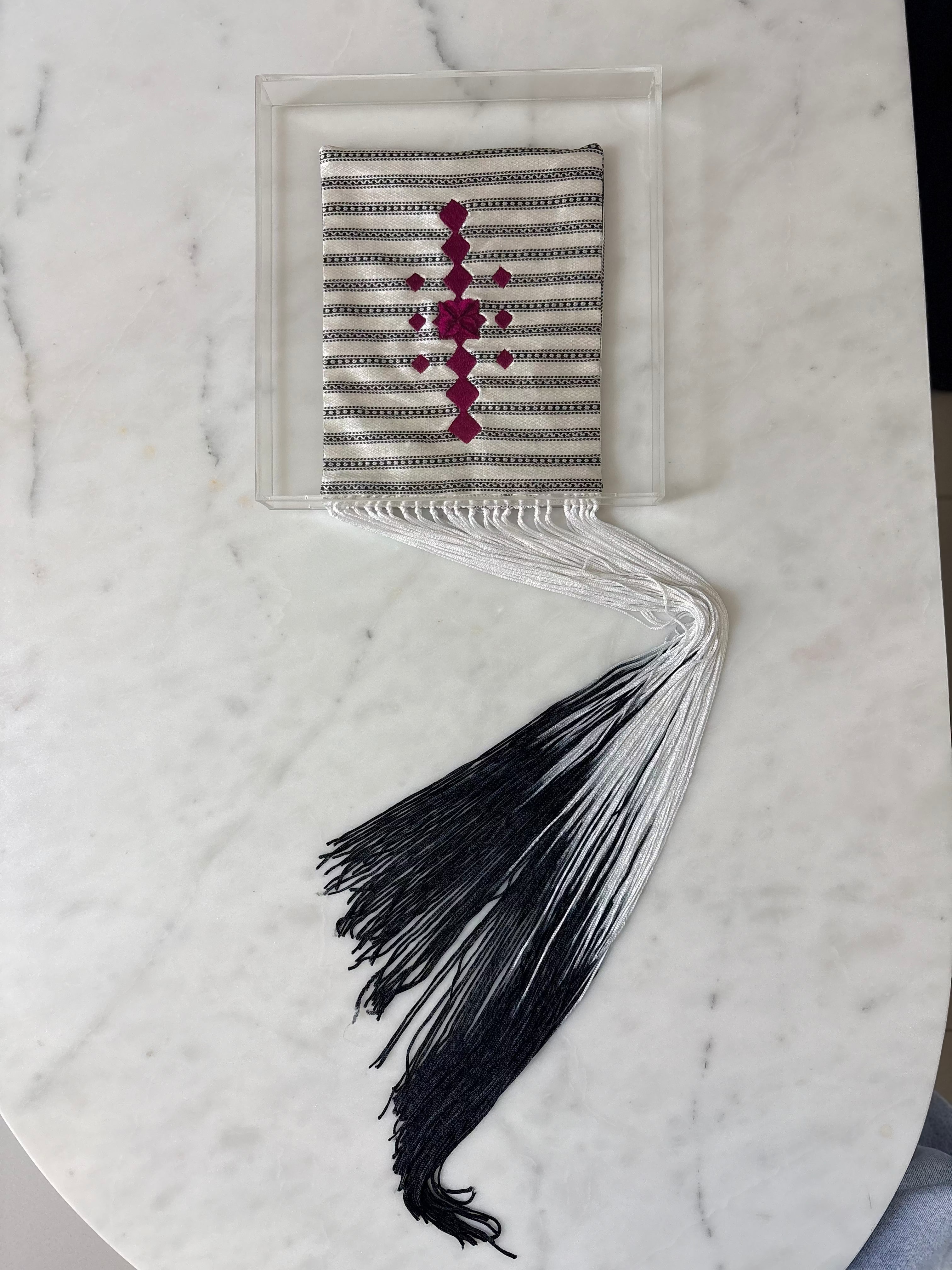 A silver and black striped shawl with a magenta embroidered cross, displayed on a white marble surface next to its black and white tassels.